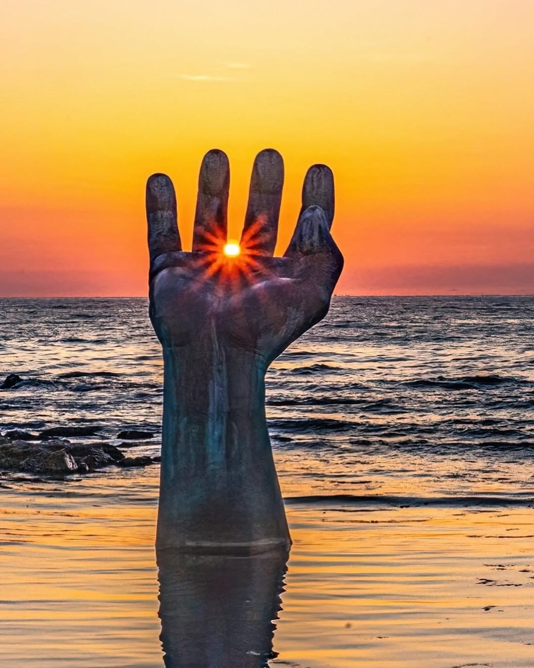 Sunrise held between the fingers of a giant hand sculpture rising from the sea